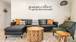Un salon moderne avec un canapé d’angle, une table basse ronde en bois et un fauteuil vert. Un mur décoratif orné d’une citation est visible.