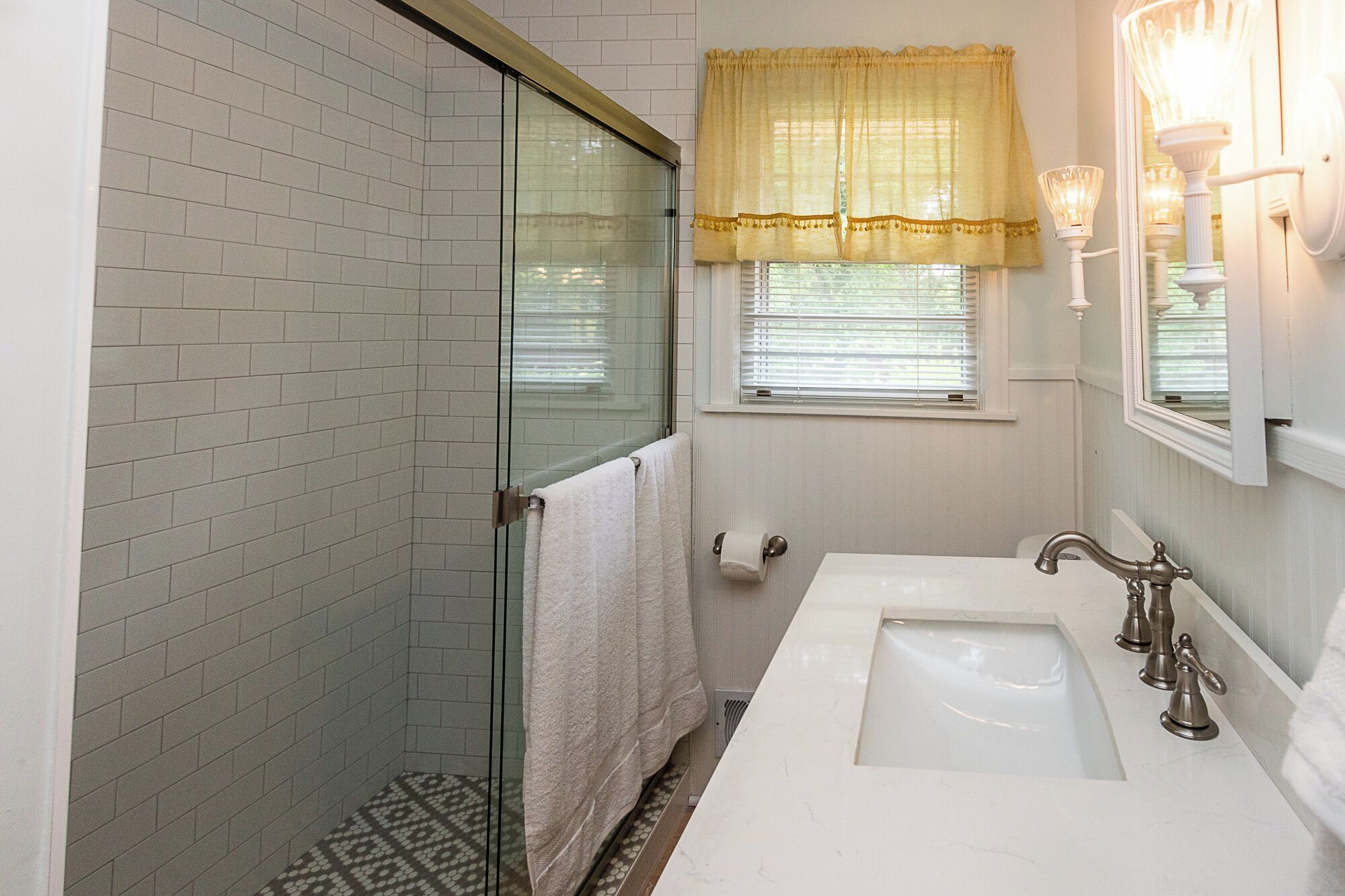 A bathroom with a glass shower door, a white sink with a vintage-style faucet, and a window with a yellow curtain.