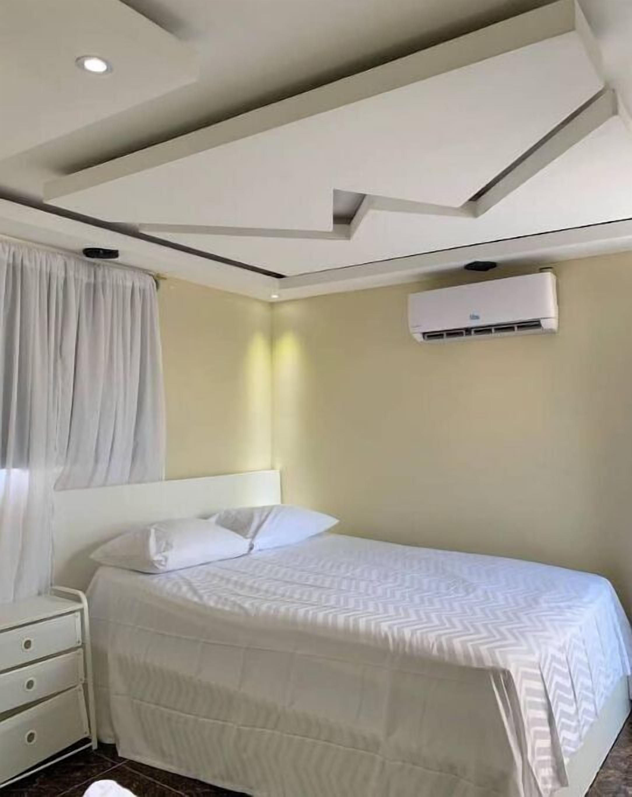 Hotellihuone, jossa on sänky, yöpöytä, seinälle asennettu ilmastointilaite ja upotetuilla valaisimilla varustettu katto.