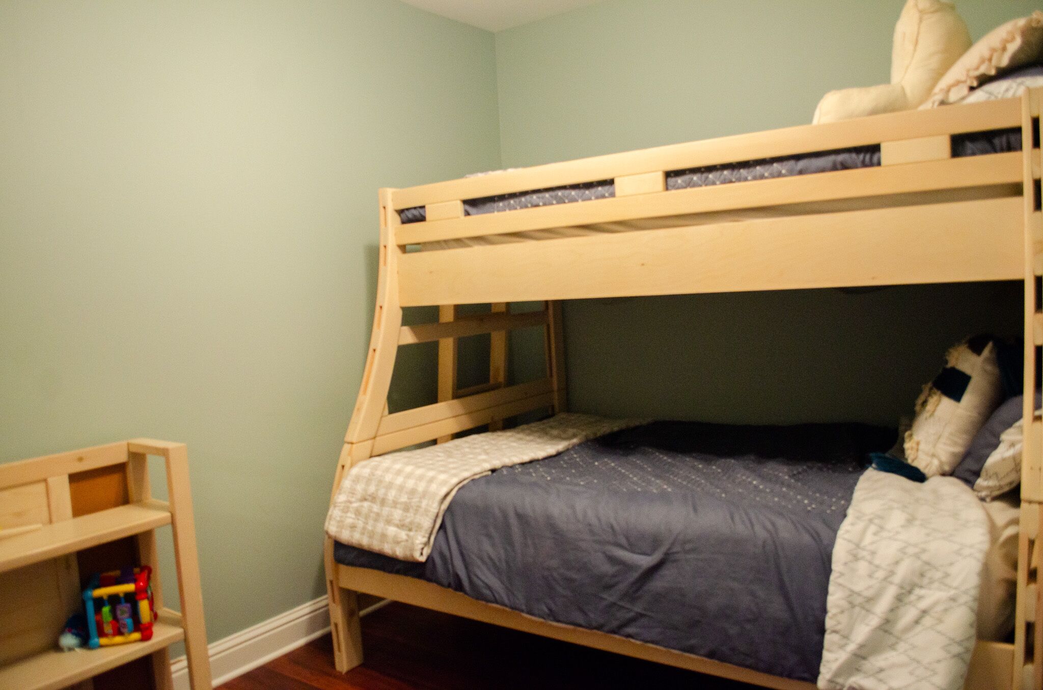 Una cama litera con colchón azul y ropa de cama blanca, una mesita de noche de madera con un juguete, y una pared de color verde claro.