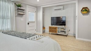 A bedroom with a bed, a TV on a wooden stand, a window with curtains, and an air conditioning unit on the wall. - Studio 1 at Beer Ranch Project. Walk to Wimberley Square. Unwind. Stay awhile. (Wimberley)