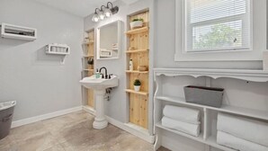 A bathroom with a pedestal sink, wooden shelving, a window with blinds, and a trash bin. - Studio 1 at Beer Ranch Project. Walk to Wimberley Square. Unwind. Stay awhile. (Wimberley)