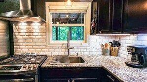 A kitchen with a gas stove, stainless steel range hood, white subway tiles, a window with a view of greenery, and a coffee maker. - Hot Tub in the Woods, Fire Pit, Pet Friendly (Eureka Springs)