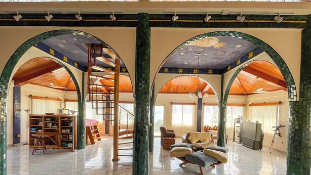 A spacious room with a high ceiling, wooden beams, and green-tiled arches. There is a ladder, a bookshelf, and a sofa. - Oceanfront Oasis with Meditation Tower & Pool (Hopeville)