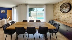 Un coin repas avec une grande table en bois et des chaises noires, un mur en pierre orné d’une horloge, et une vue sur une piscine à travers des portes vitrées.