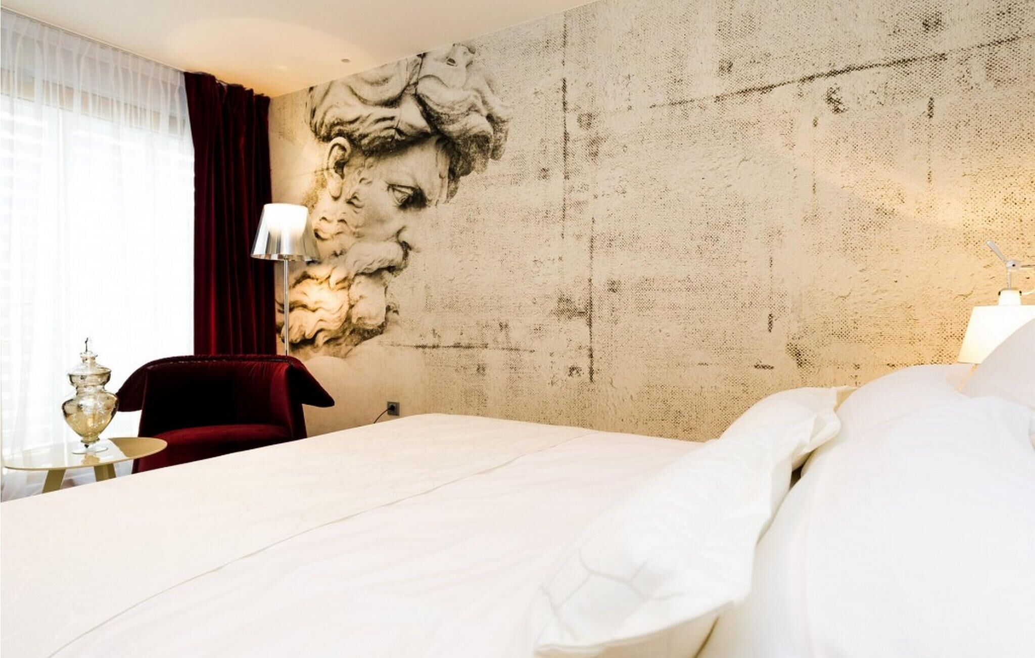 A hotel room with a large bed, a red chair, a lamp, and a wall with a textured mural.