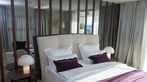 A hotel room with a large bed, two bedside tables with lamps, and a view of the sea through the window.