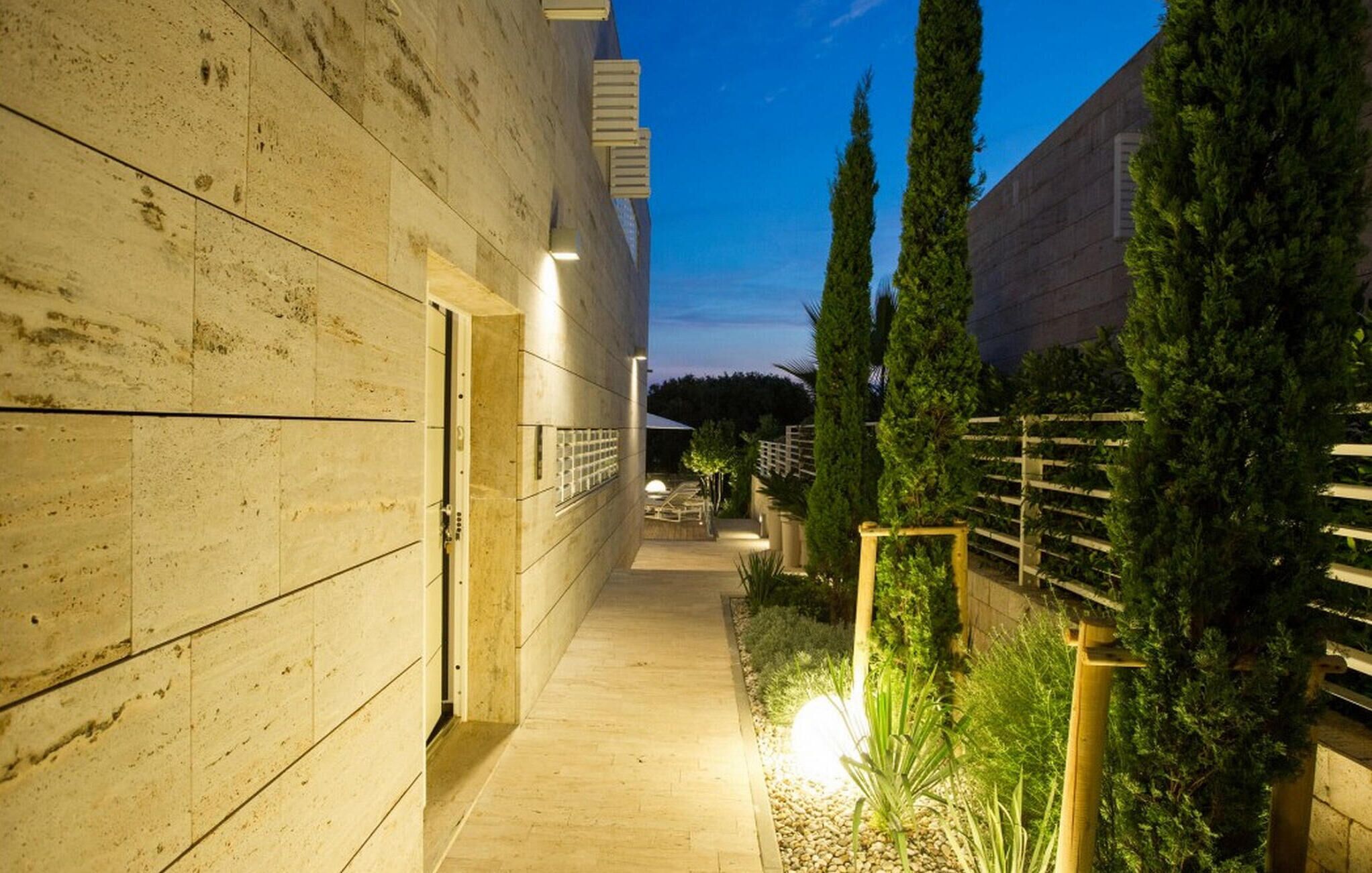 Un sendero estrecho que lleva a un edificio con fachada de madera y un pequeño jardín con árboles altos y una valla de madera.