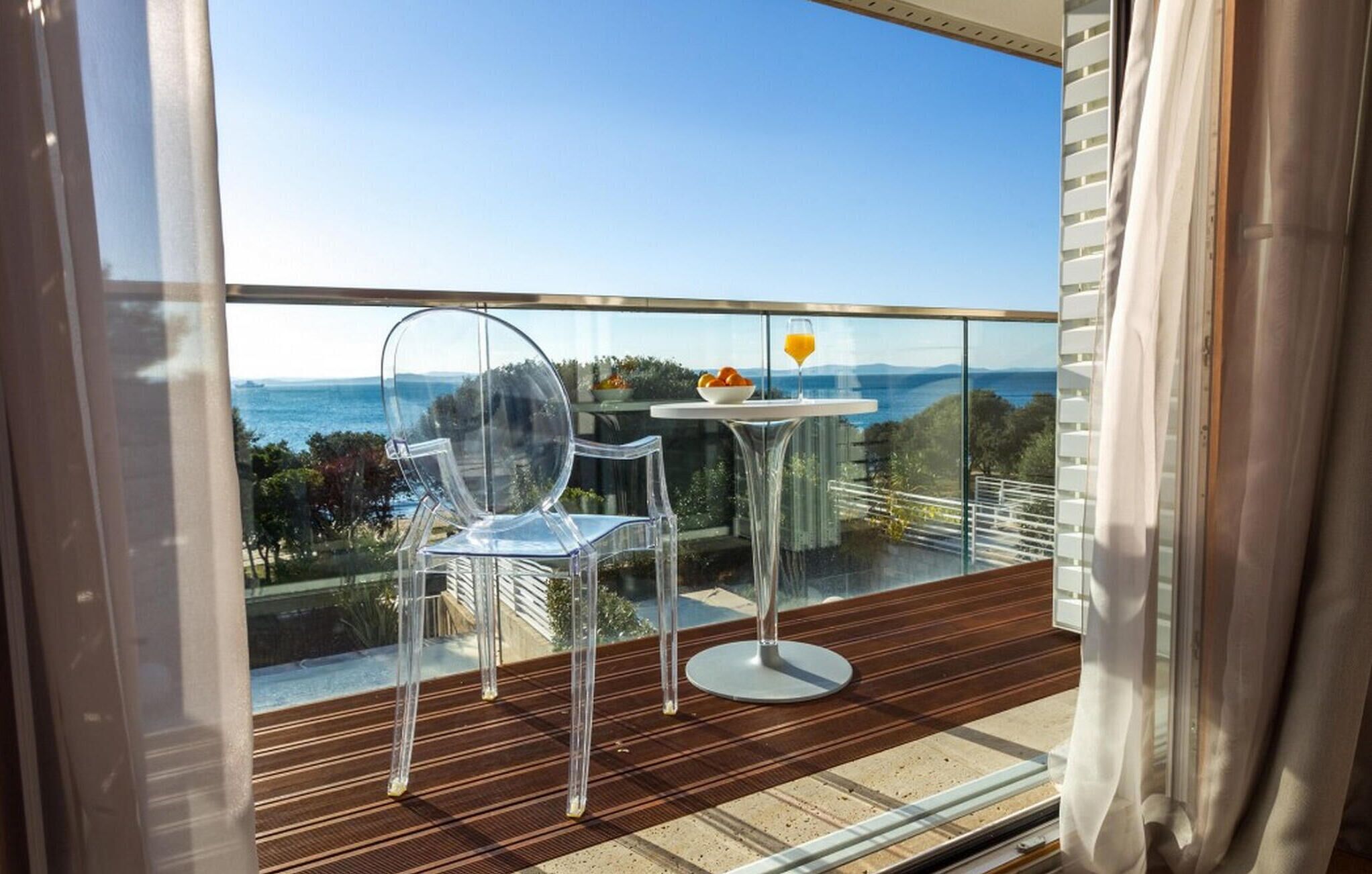 A balcony with a clear view of the sea, two transparent chairs, a small table with a bowl of fruit and a glass of orange juice, and a wooden floor.
