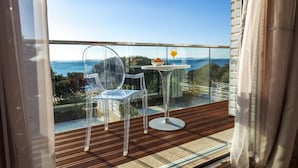 A balcony with a clear view of the sea, two transparent chairs, a small table with a bowl of fruit and a glass of orange juice, and a wooden floor.