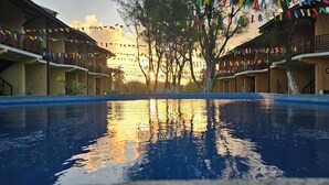 A swimming pool with a sunset reflection, surrounded by hotel buildings with balconies and colorful flags. - Condomínio Frente mar pé na Área, Piscinas Naturais! Casa Térreo Aconchegante (Camaçari)