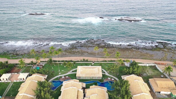 An aerial view of a resort with a beachfront, a swimming pool, and multiple bungalow-style buildings. - Condomínio Frente mar pé na Área, Piscinas Naturais! Casa Térreo Aconchegante (Camaçari)