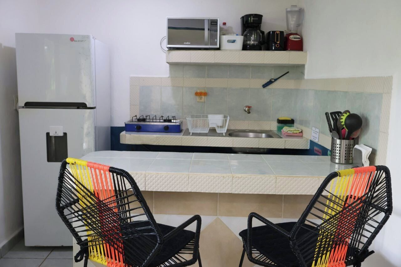 Cocina compacta con refrigerador, microondas y estufa a gas. Hay dos sillas con respaldos de colores y una pequeña zona de lavamanos.