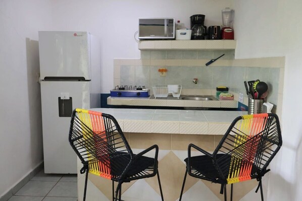 A compact kitchen with a refrigerator, microwave, stove, and a small sink. There are two chairs with a colorful backrest.