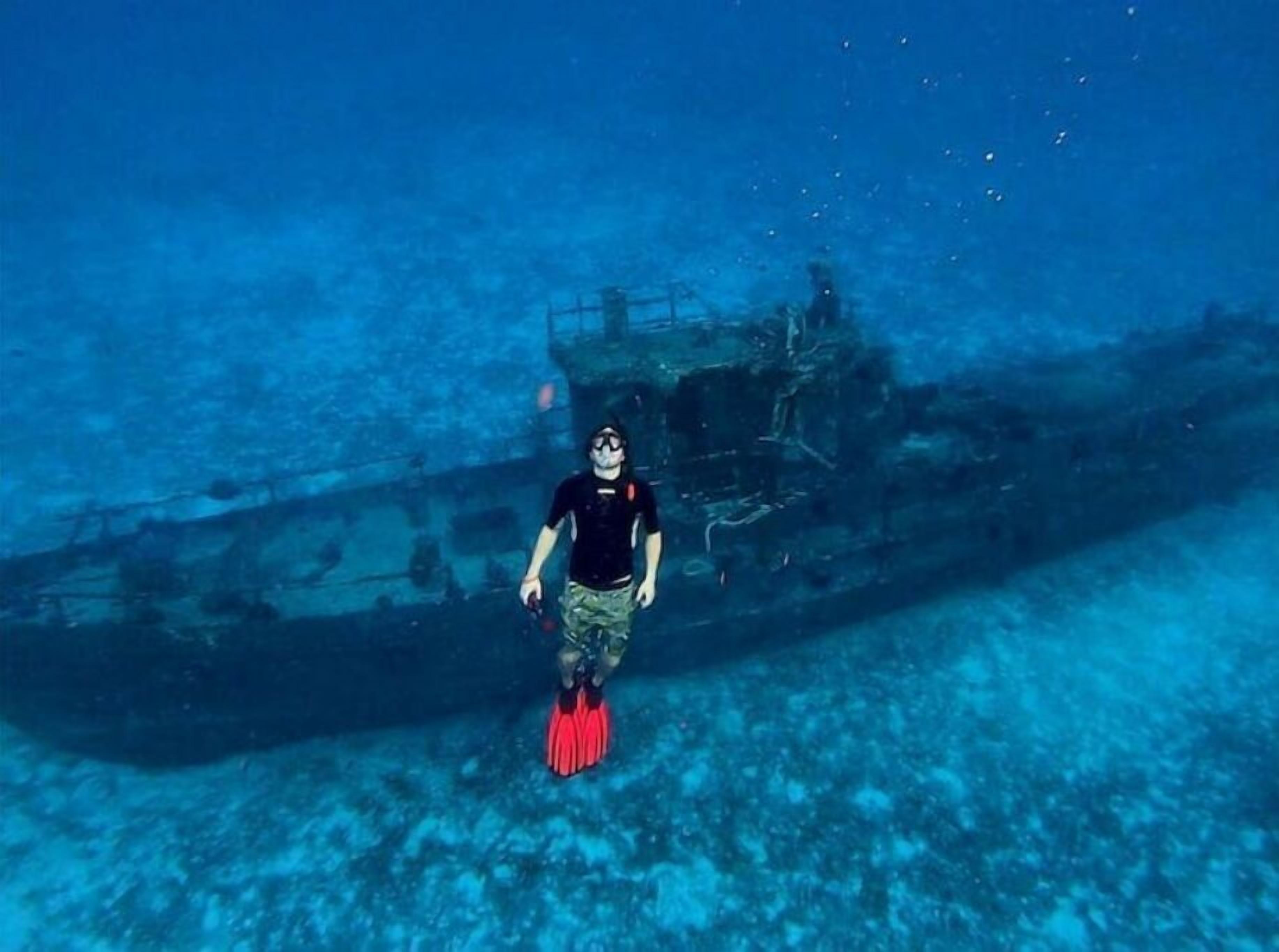 A diver in a black shirt and camouflage shorts with red fins, swimming near a sunken shipwreck.