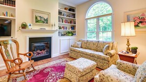 A cozy living room with a fireplace, floral-patterned furniture, a bookshelf, and a large window with a view of greenery. - Charming Flushing Cottage w/ Deck + Spacious Yard! (Flushing)