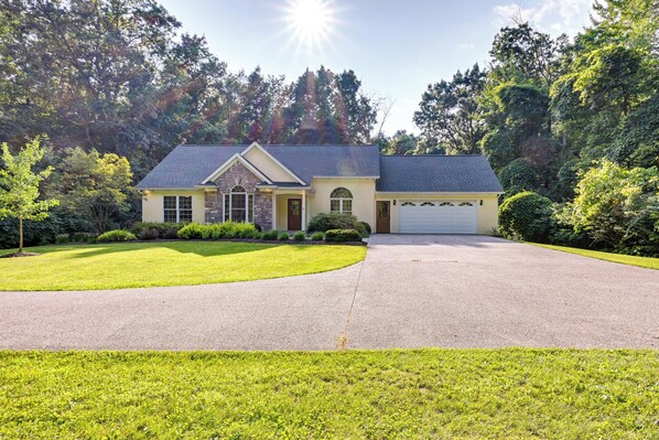 A single-story house with a garage, surrounded by a well-maintained lawn and dense trees. - Charming Flushing Cottage w/ Deck + Spacious Yard! (Flushing)