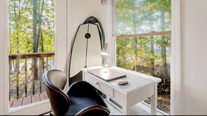 A room with a desk, chair, surfboard, and large windows overlooking a forest. - Lazy Creek Cove Cottage w EV Plug! (Chapin)