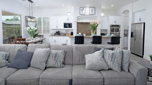 A modern living room with a grey sectional sofa, a dining area with a table and chairs, and a kitchen with white cabinets and stainless steel appliances. - Luxurious 5BR Resort Style Home w/ Pool & Spa (Indio)