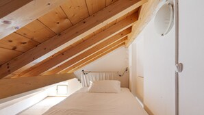 Une chambre avec un plafond en pente, un lit, une fenêtre et un ventilateur fixé au mur.