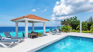 A poolside area with lounge chairs, a covered seating area, and a view of the ocean. - The Beach House - Coral Estate Resort (Sint Willibrordus)