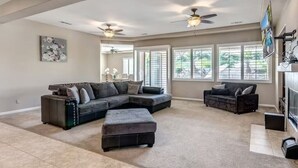 A spacious living room with a sectional sofa, ottoman, ceiling fans, large windows, and a flat-screen TV. - Lux Desert Oasis w/ saltwater pool near Coachella (Indio)