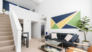 A modern living room with a staircase, a glass coffee table, a sofa, and a large abstract painting on the wall.