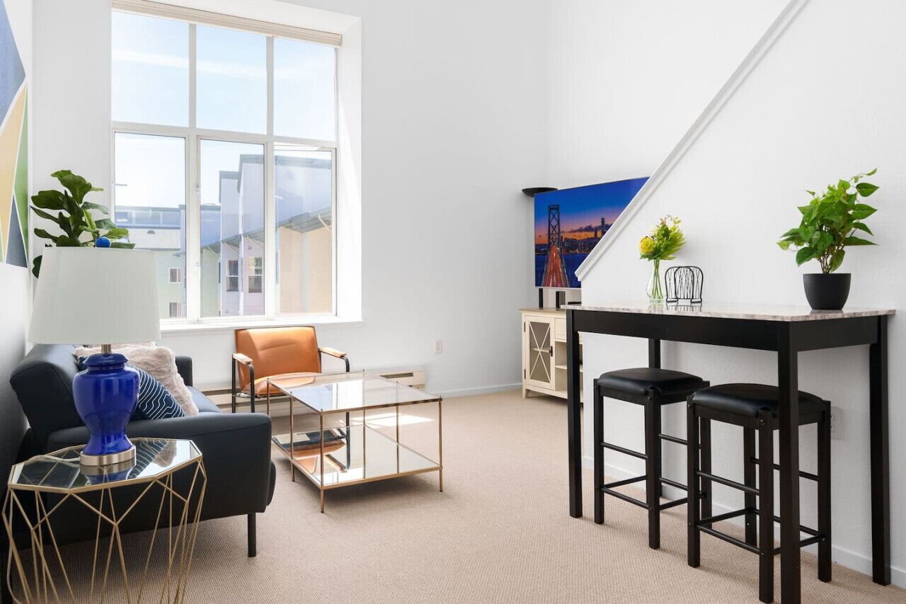 Una sala moderna con una mesa de centro de vidrio, un sofá negro, una mesa de comedor negra con dos taburetes y un cuadro de un paisaje urbano en la pared.