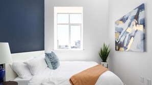 A bedroom with a bed, a window, a plant, and a painting.