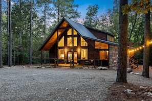 A log cabin with a stone foundation, large windows, and a porch with outdoor seating. - Secluded Creekside Cabin | Hot Tub | Dog Friendly (Broken Bow)
