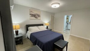 Een slaapkamer met een bed, nachtkastjes en een nachtkast. Er hangt een schilderij aan de muur en een deur die leidt naar een andere kamer.
