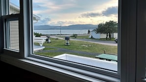Vista desde una ventana a un área verde con una parrilla y una mesa de picnic, con vista a un cuerpo de agua y montañas distantes.