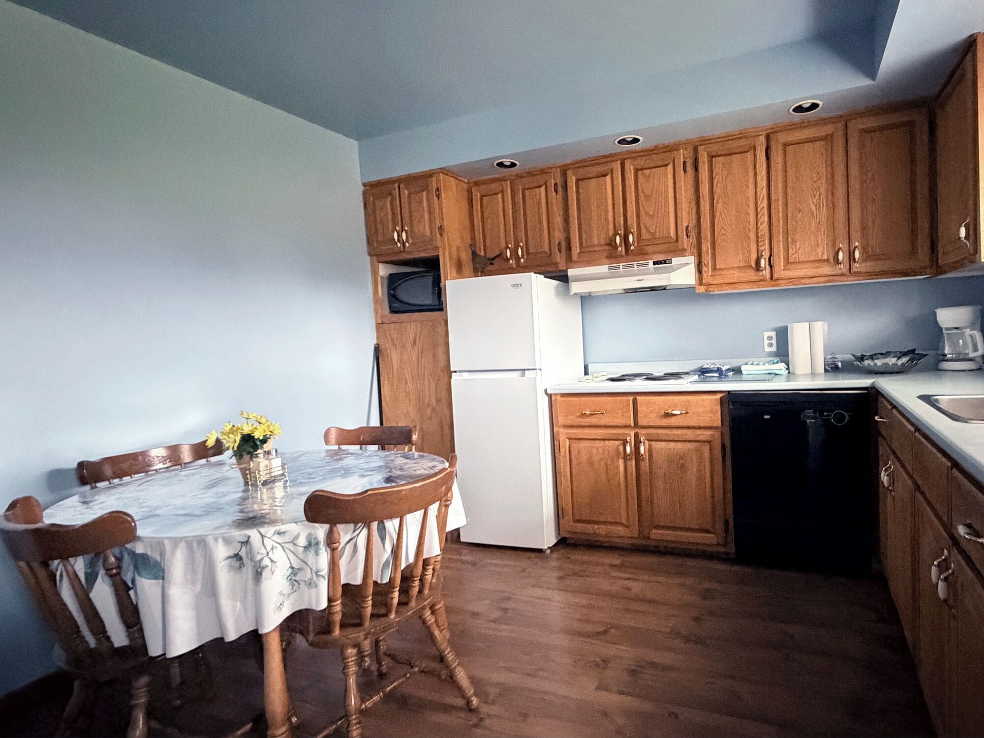 A compact kitchen with wooden cabinets, a white refrigerator, a microwave, and a dishwasher. There is a round table with chairs and a vase with flowers.