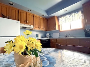 Una cocina con armarios de madera, un refrigerador, una cafetera y un florero con flores amarillas sobre la mesa.