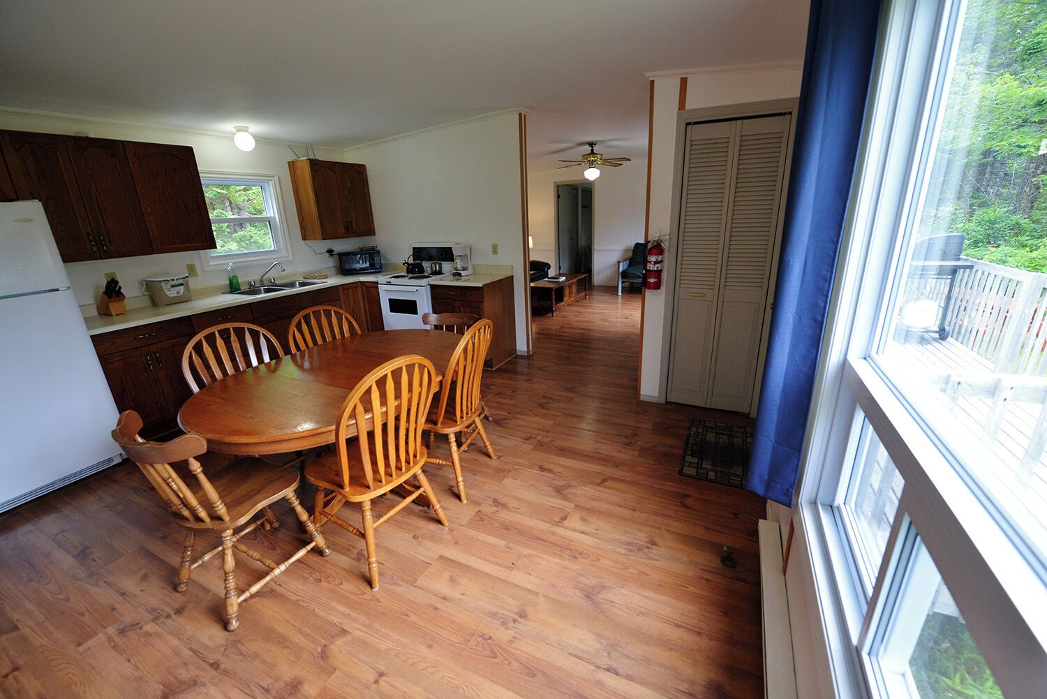 A kitchen with wooden cabinets, a dining table with chairs, a refrigerator, and a window with a view of greenery.