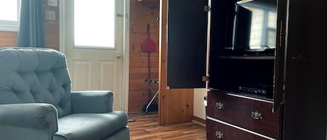 Una stanza con una poltrona trapuntata, un pavimento in legno, un armadio con cassetti, una porta e una finestra con tende.