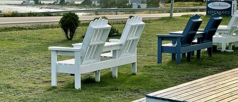 Outdoor seating with Adirondack chairs and a wooden deck, overlooking a body of water and distant buildings.