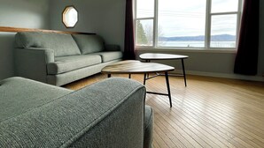 Una habitación con un sofá gris, una mesita redonda pequeña y un ventanal con cortinas.