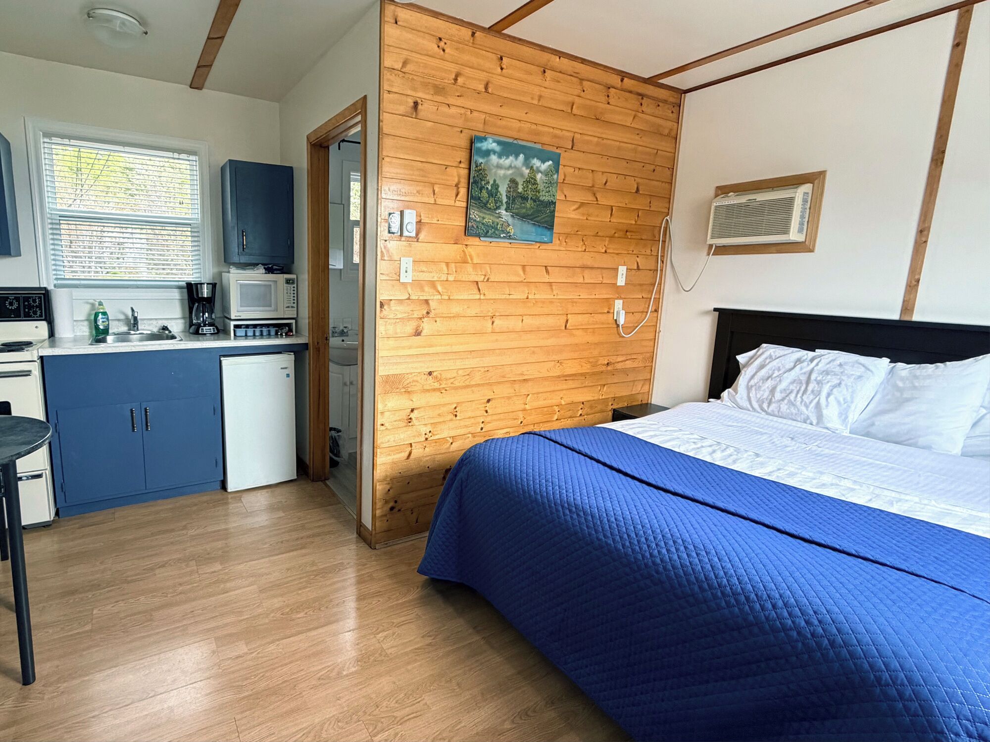 A bedroom with a bed, a wooden wall, a painting, and a kitchenette with a microwave and cabinets.