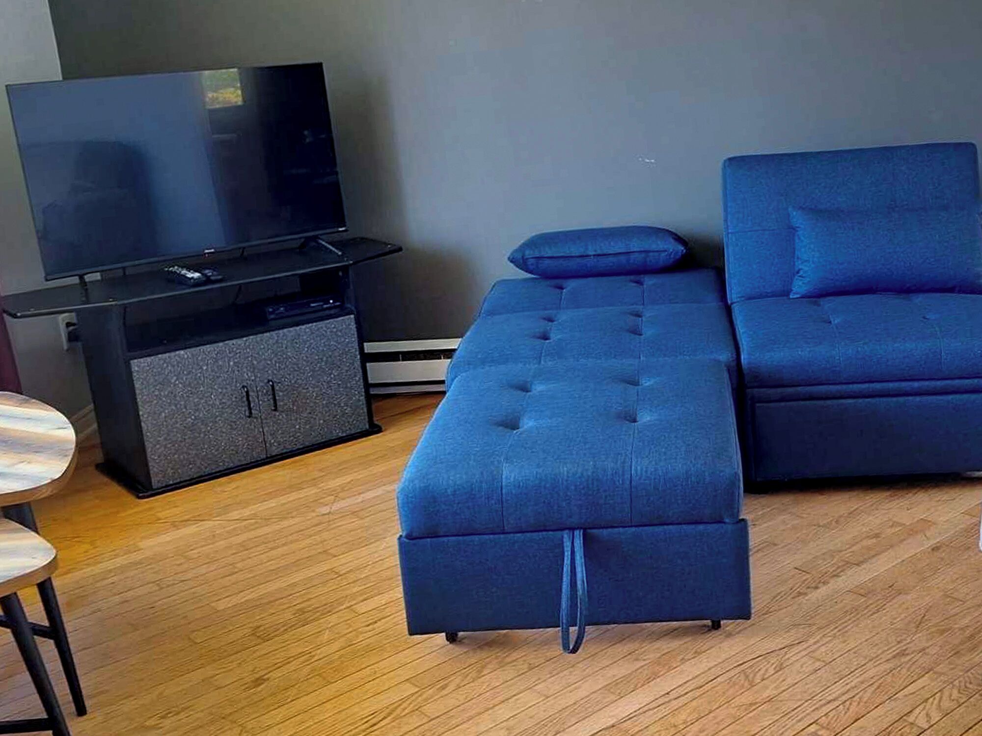 A modern living room with a flat-screen TV, a dark blue sofa, a matching ottoman, and a small wooden table.