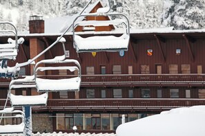 A ski lift chair covered in snow, a wooden building with multiple windows, and a snowy mountain in the background.