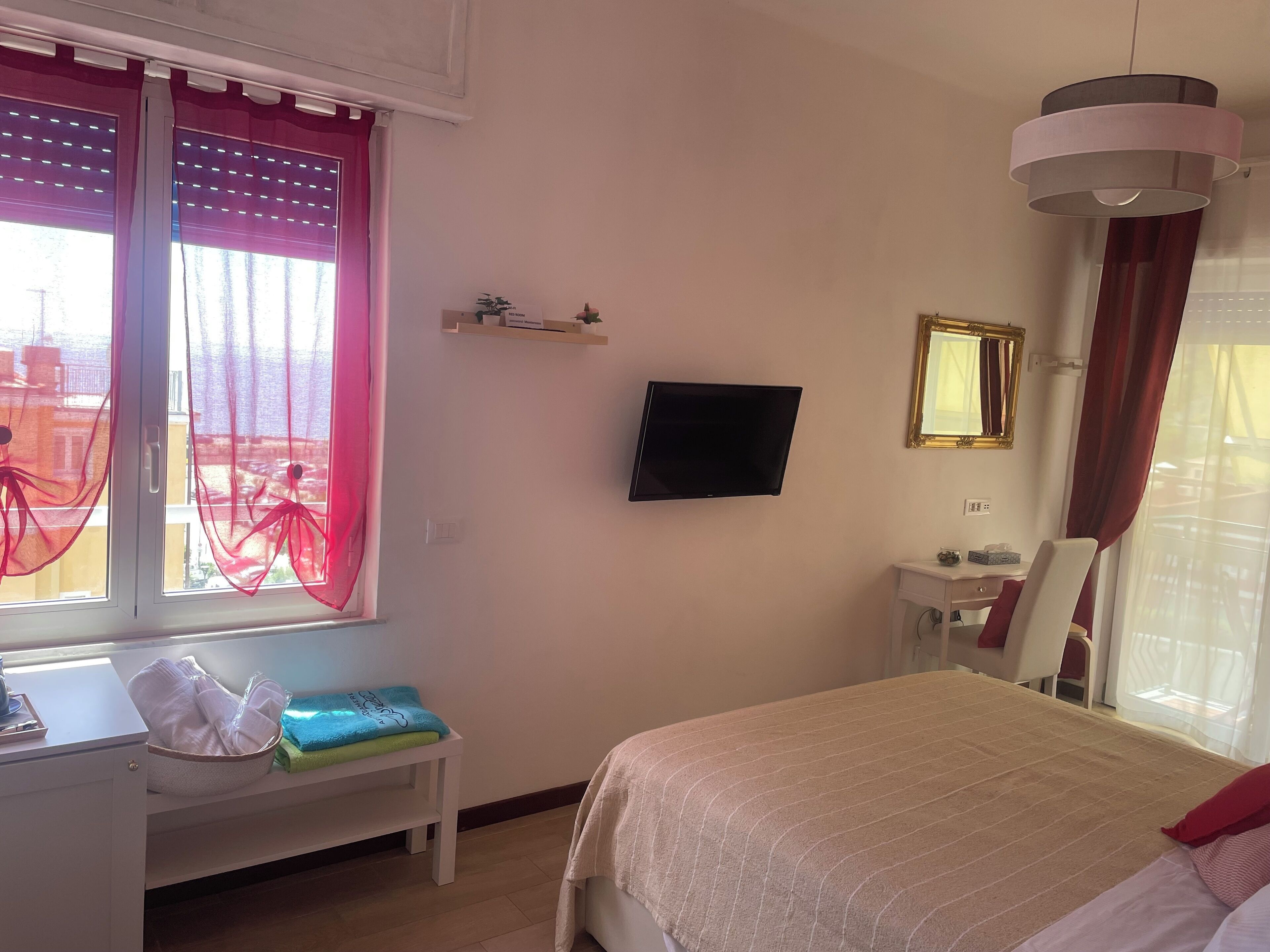 A hotel room with a bed, a desk with a chair, a television, a small shelf with items, and a window with curtains.