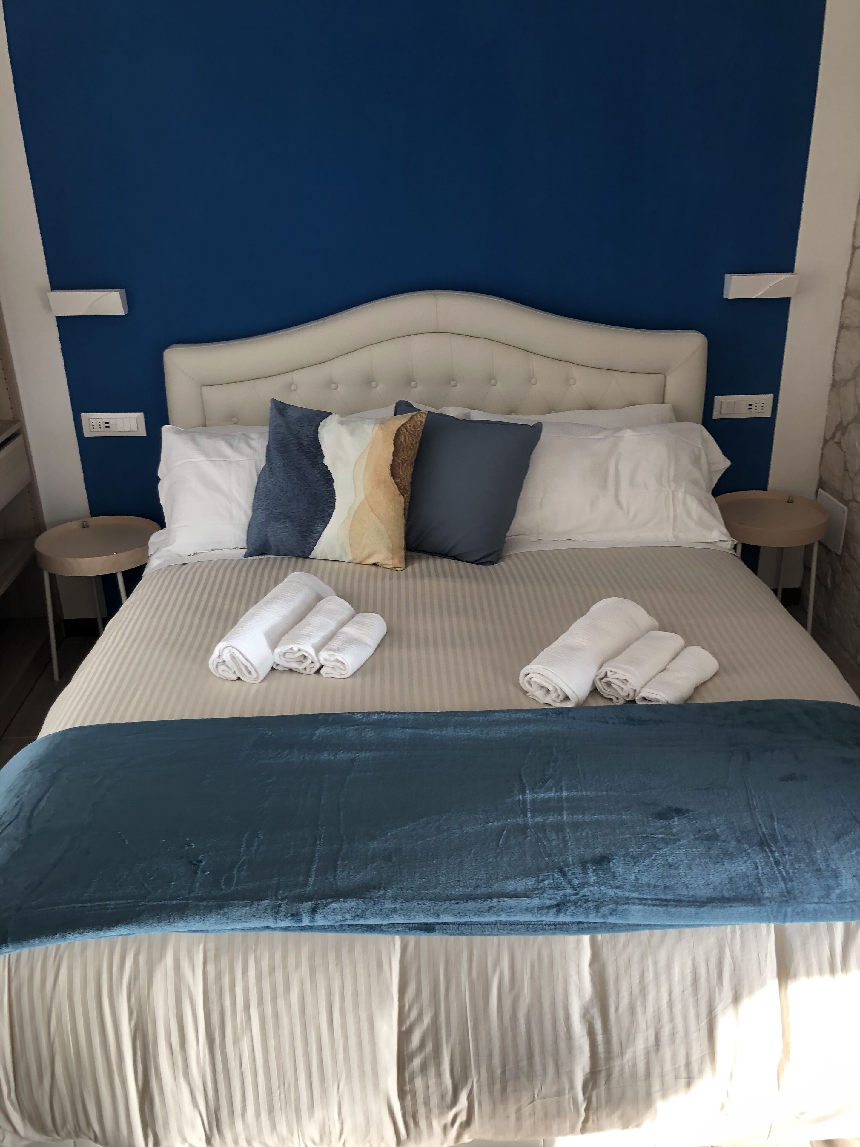 A neatly made bed with a blue blanket, white pillows, and rolled towels.