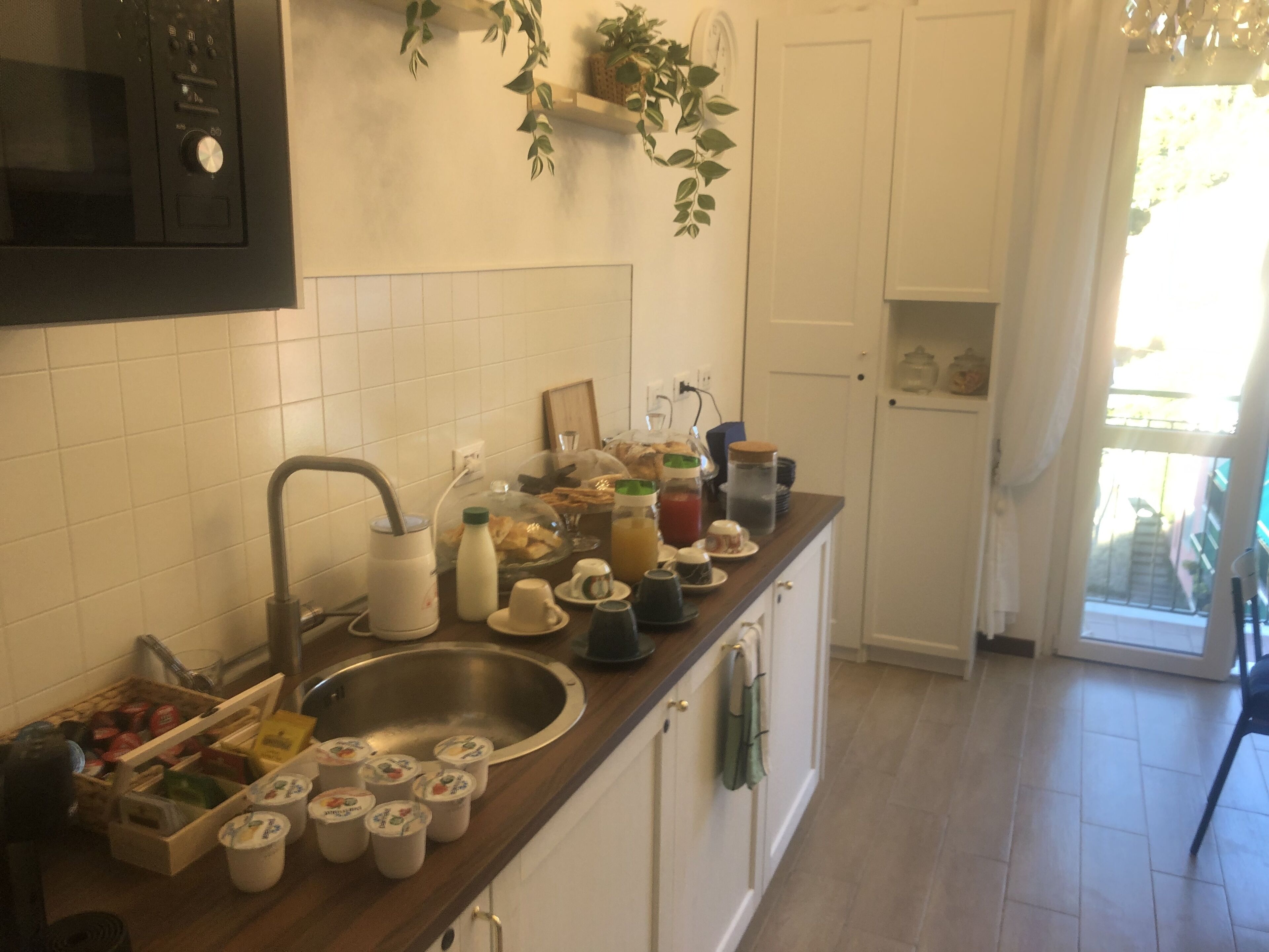 Eine Küche mit einer Edelstahl-Spüle, weißen Schränken und einer Mikrowelle. Auf der Arbeitsplatte befinden sich verschiedene Küchenutensilien, darunter eine Kaffeemaschine, Tassen und Gläser.