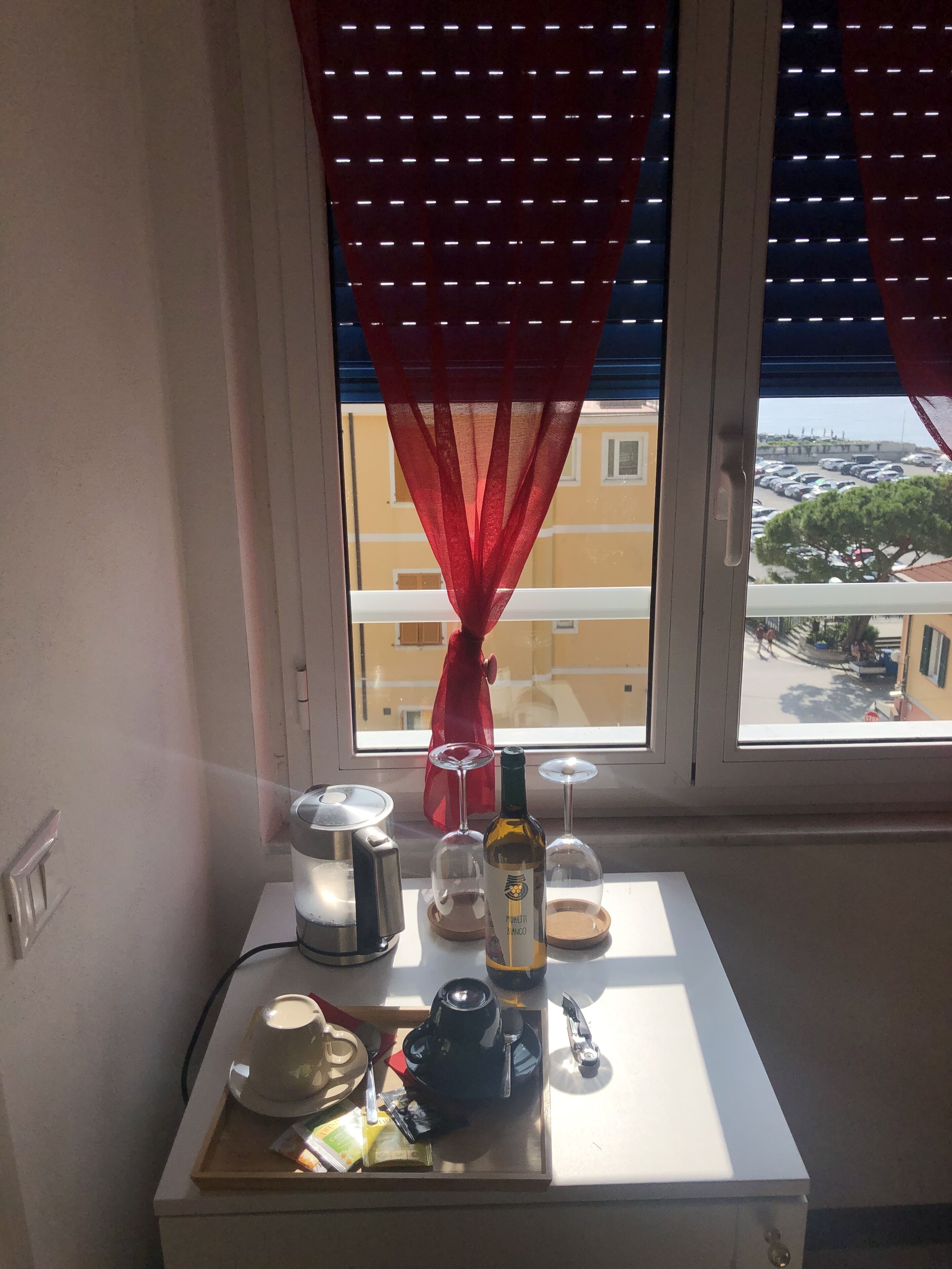Ett fönster med röda gardiner, ett bord med en kaffebryggare, en flaska och ett bricka med diverse föremål.
