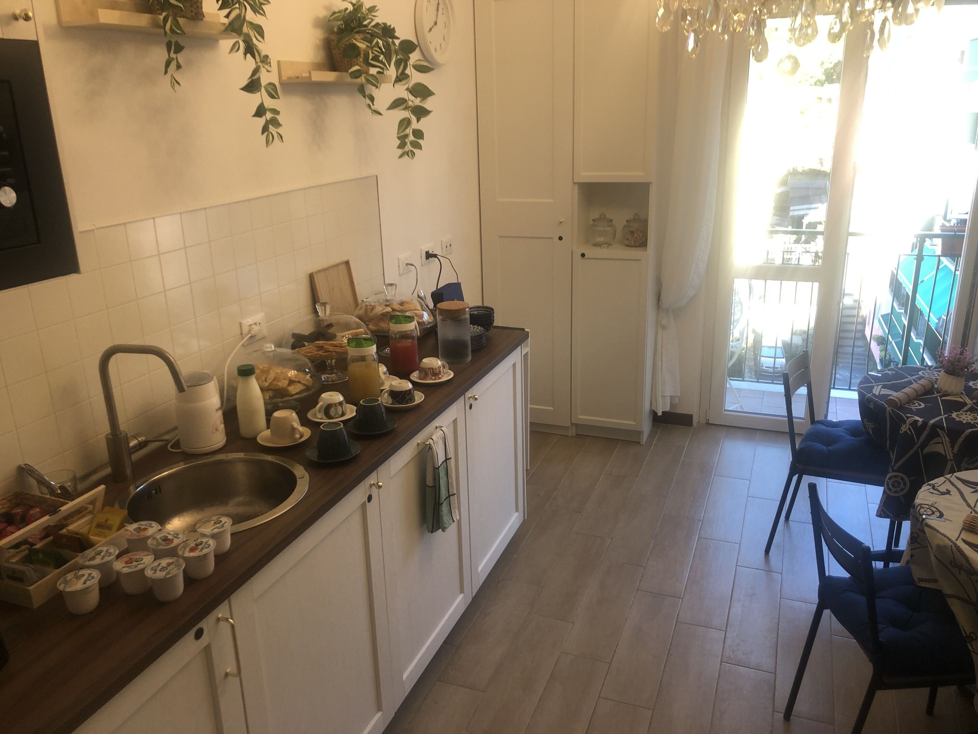 Une cuisine avec des armoires blanches, un plan de travail en bois et un évier. On peut voir divers ustensiles de cuisine sur le plan de travail, notamment des tasses, des bocaux et une bouilloire. Une plante est suspendue au mur au-dessus du plan de travail, et une fenêtre avec des rideaux est visible à l’arrière-plan.