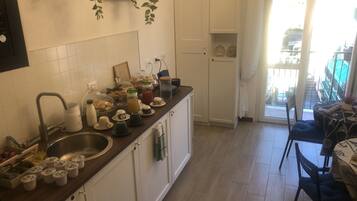 Une cuisine avec des armoires blanches, un plan de travail en bois et un évier. On peut voir divers ustensiles de cuisine sur le plan de travail, notamment des tasses, des bocaux et une bouilloire. Une plante est suspendue au mur au-dessus du plan de travail, et une fenêtre avec des rideaux est visible à l’arrière-plan.