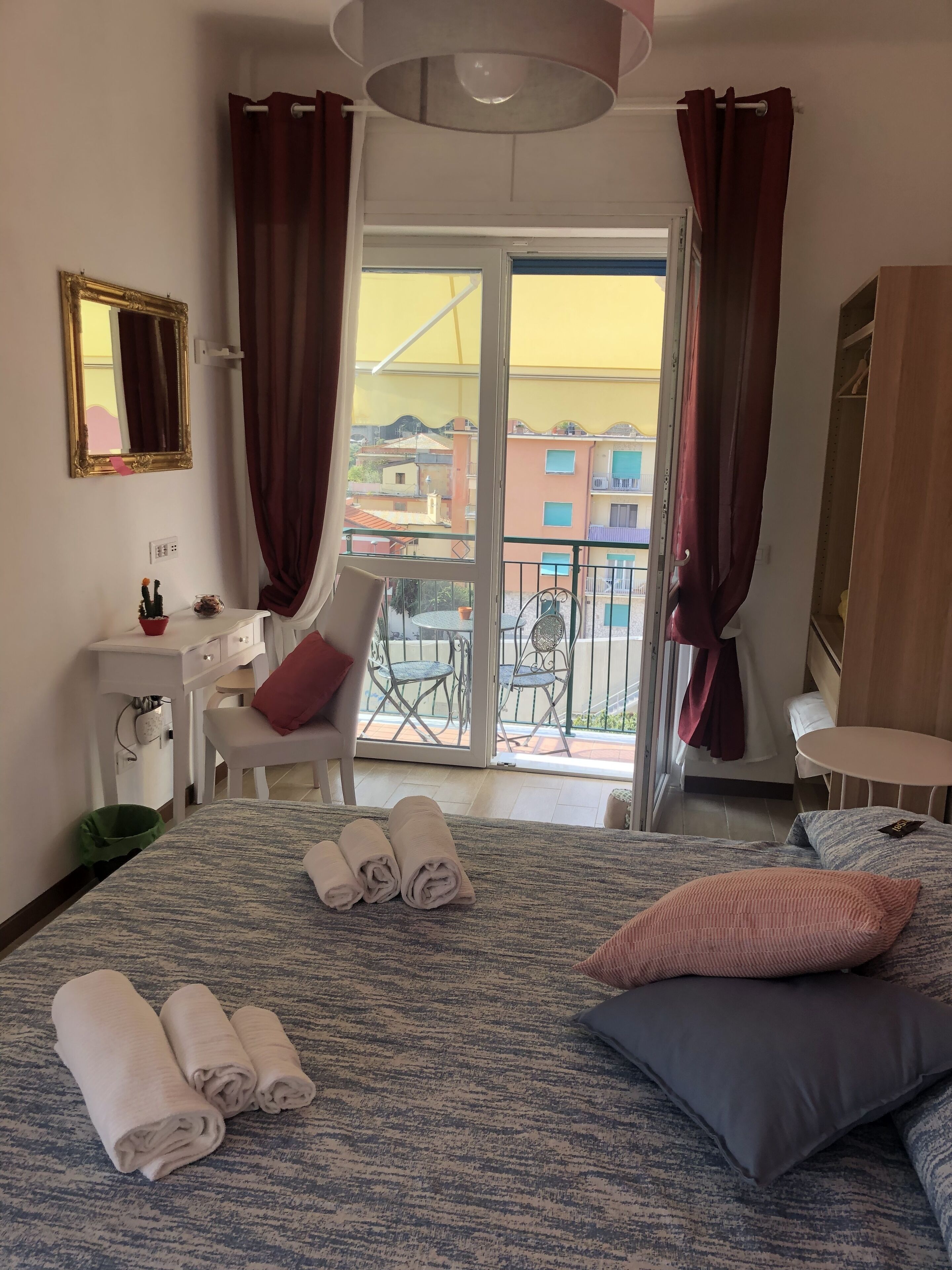 A hotel room with a bed, a desk, a chair, a balcony with a view of buildings, and a large mirror on the wall.