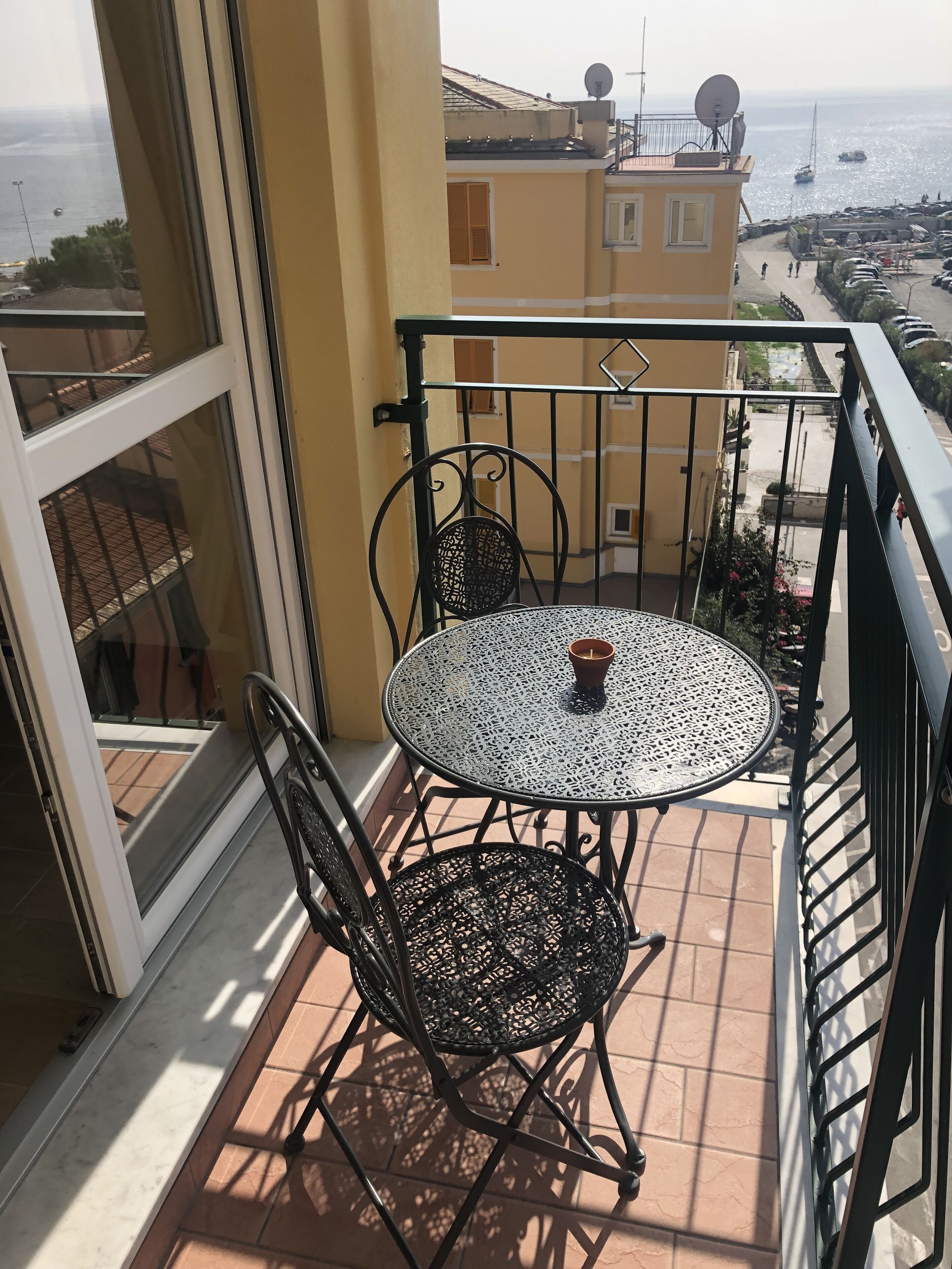 A balcony with a round table and two chairs, overlooking a coastal area with buildings and boats.