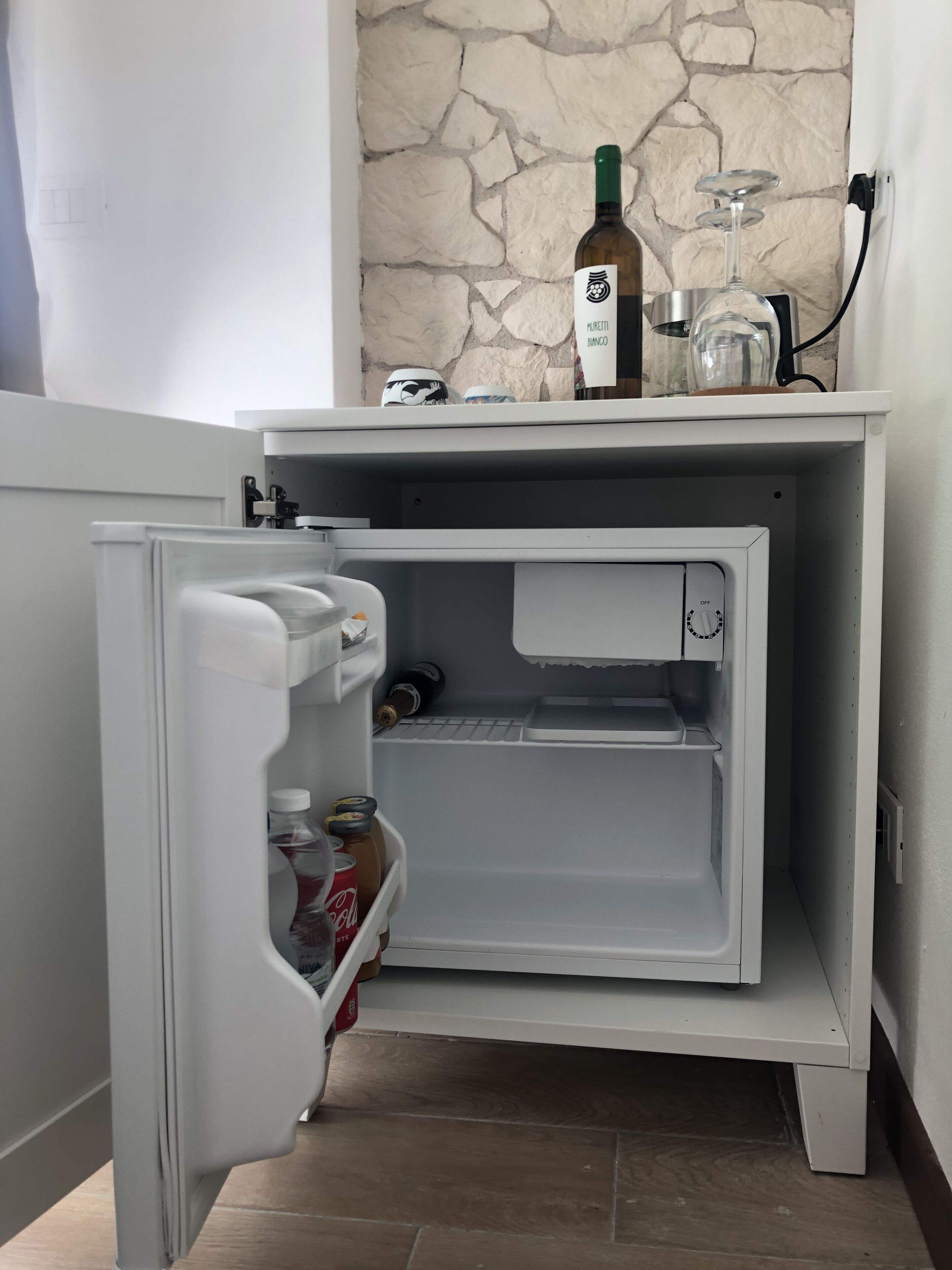 Un petit réfrigérateur, avec un mur en pierre en arrière-plan, une bouteille de vin et un verre à vin posés sur le dessus.